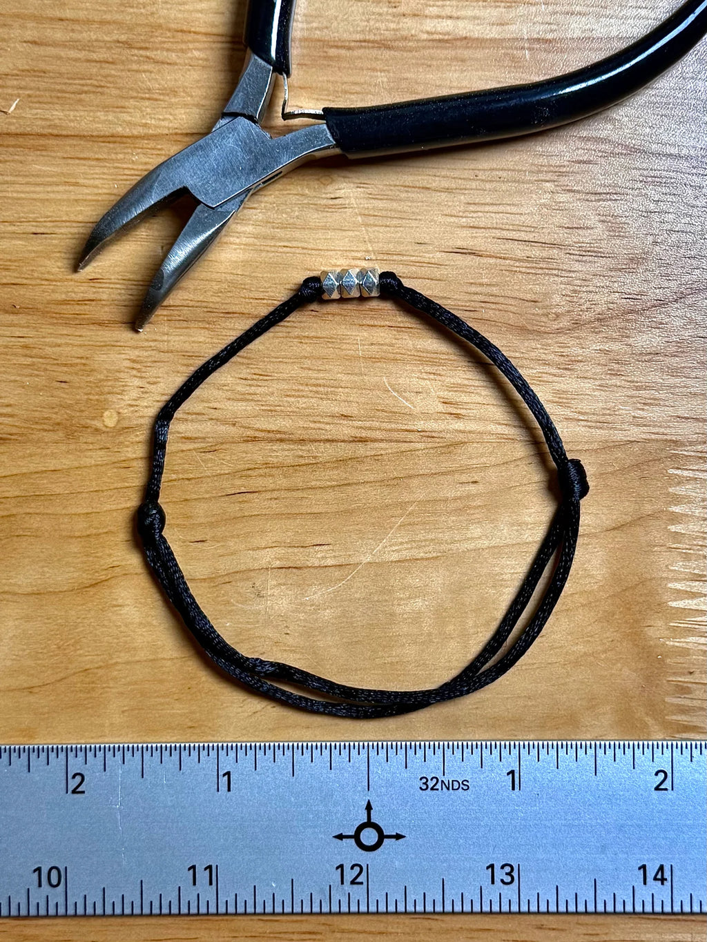 A black adjustable cord bracelet with three silver spacer beads, displayed on a wooden surface next to a metal ruler and a pair of pliers.