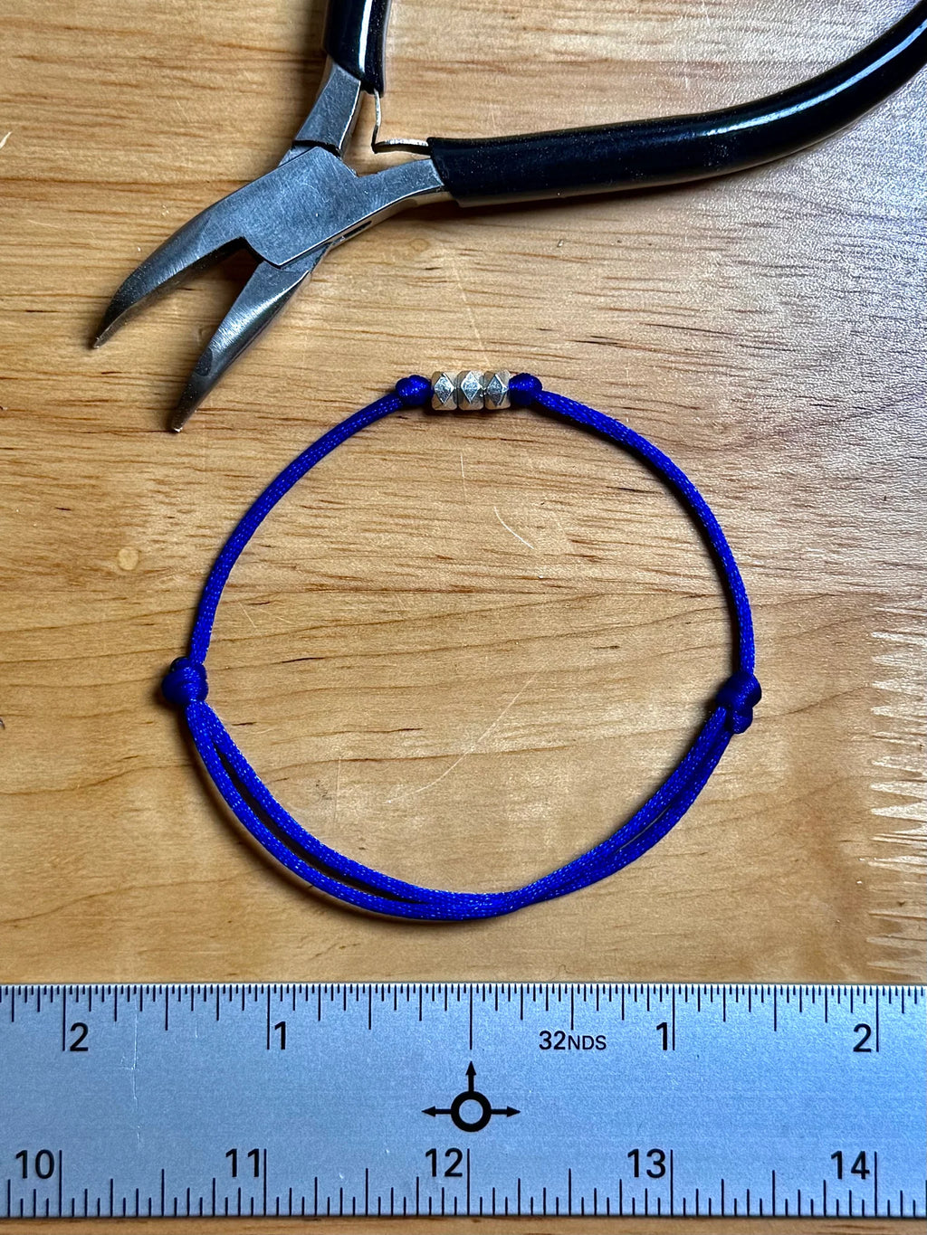 A blue adjustable cord bracelet with three silver spacer beads, displayed on a wooden surface next to a metal ruler and a pair of pliers.