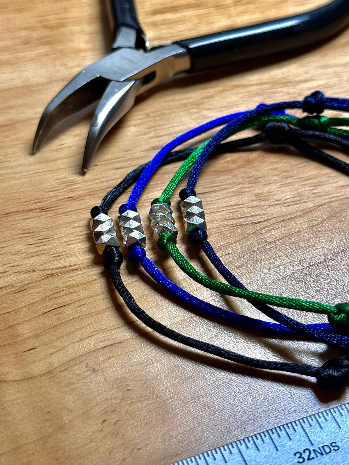 Four colorful bracelets with silver beads on a wooden surface with pliers and a ruler.