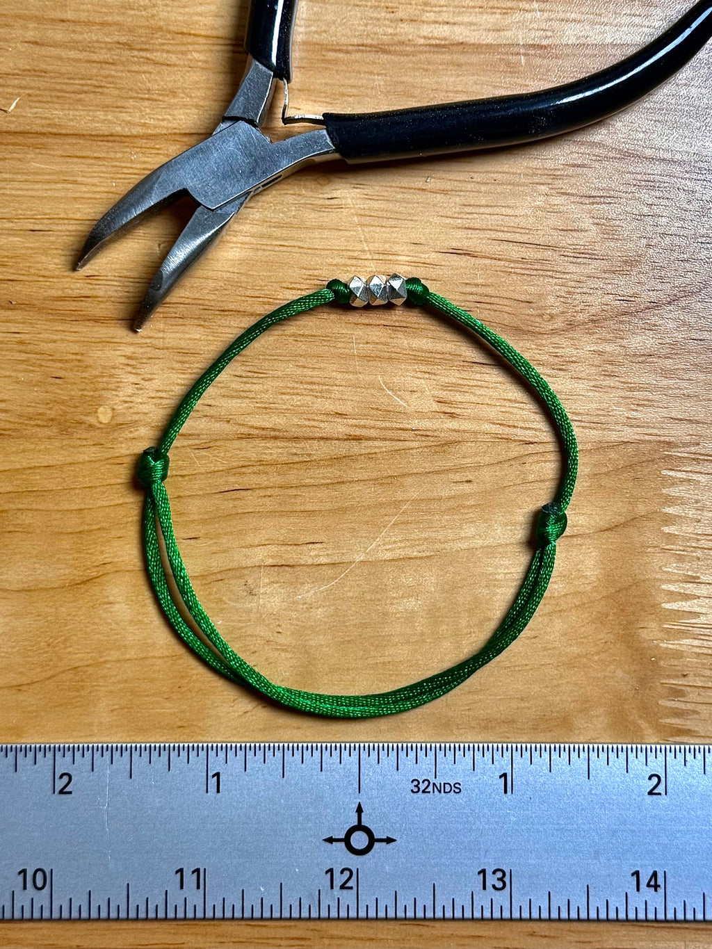 A green adjustable cord bracelet with three silver spacer beads, displayed on a wooden surface next to a metal ruler and a pair of pliers.
