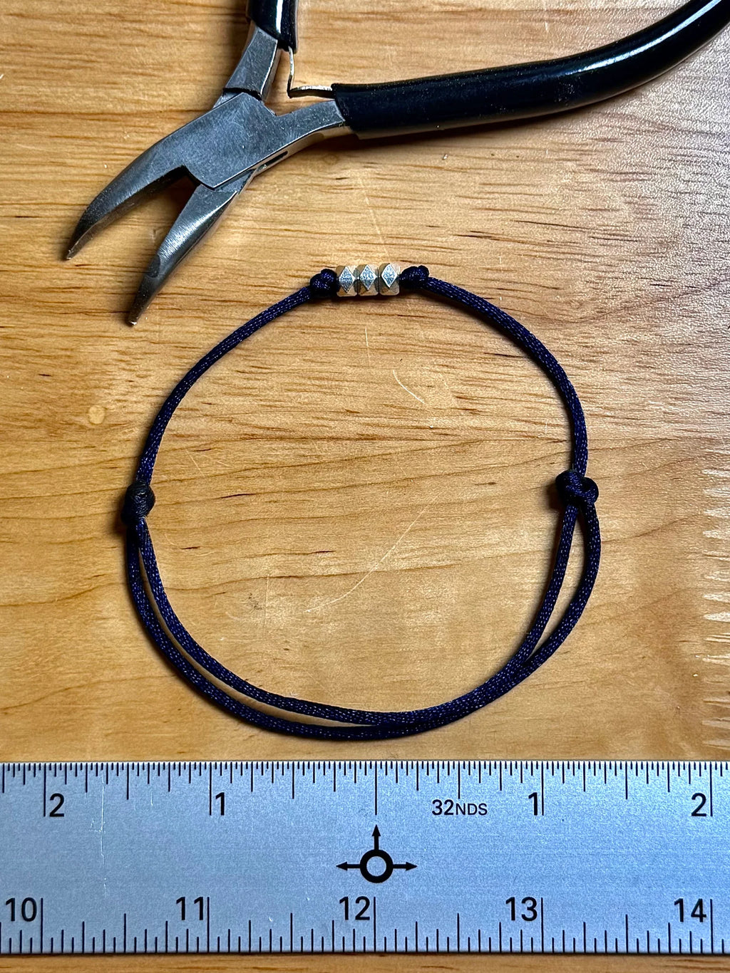 A navy blue adjustable cord bracelet with three silver spacer beads, displayed on a wooden surface next to a metal ruler and a pair of pliers.